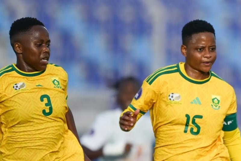 Two female soccer players in yellow South Africa jerseys (numbers 3 and 15) run on the field during a match.