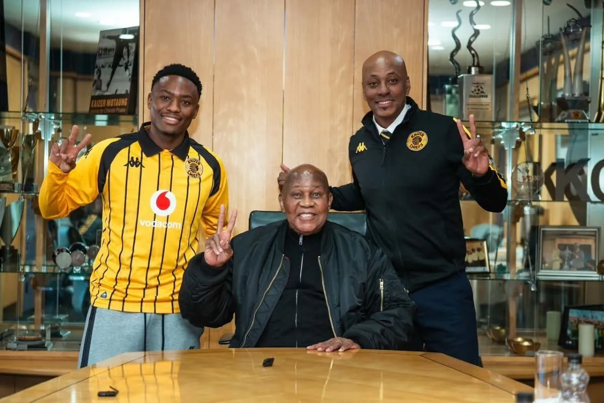 Three men posing in an office, seated center with two teammates standing on either side, all smiling and flashing peace signs, trophies in the background.