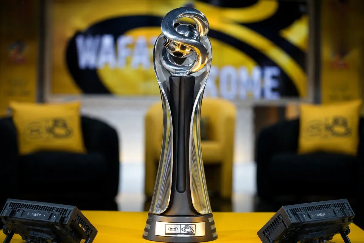 Silver trophy with twisting metal arms on a black base sits on a yellow table in a blurred event setting with lounge chairs in the background.